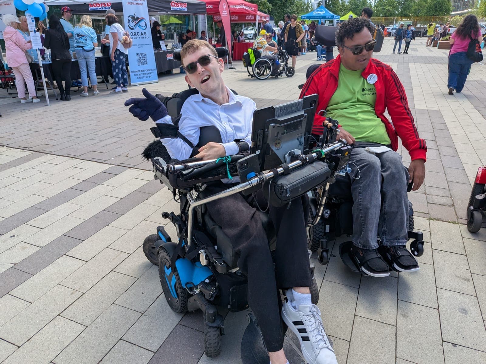 Two men in motorized wheelchairs smile and point at an outdoor event with vendor tents.