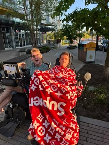 Two people in motorized wheelchairs outdoors, one covered in a red Supreme and Louis Vuitton blanket.