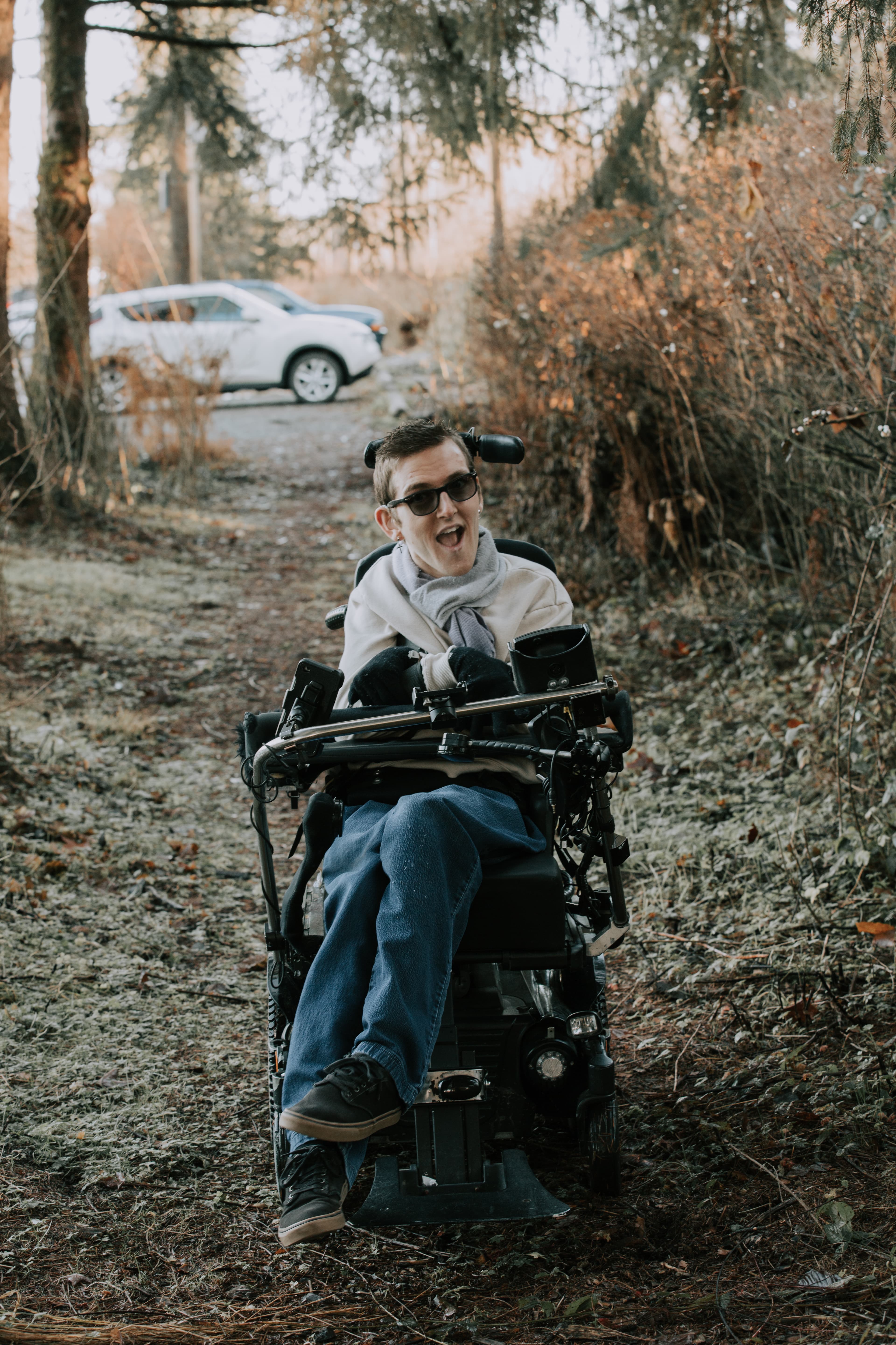 Smiling person in a motorized wheelchair wearing sunglasses navigates a wooded trail outdoors.